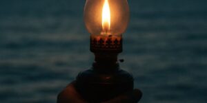 Eine Hand hält eine Lampe mit einer brenndenden Kerze und im Hintergrund ist das Meer zu sehen